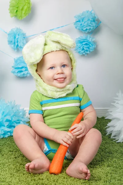 Retrato de lindo adorable bebé caucásico en vacaciones traje de conejo de Pascua con orejas grandes, vestido con ropa verde onesie, sentado en la alfombra suave alfombra esponjosa en el estudio, sosteniendo zanahoria roja — Foto de Stock