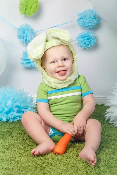 Portrait of cute adorable Caucasian baby boy in holiday Easter bunny rabbit costume with large ears,  dressed in green clothes onesie, sitting on soft fluffy rug carpet in studio, holding red carrot — Stock Photo, Image