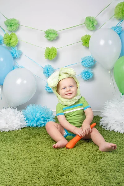 Porträtt av söt bedårande kaukasiska baby boy i semester påsk kanin kostym med stora öron, klädd i gröna kläder onesie, sitter på mjuk fluffig matta matta i studio, hålla röd morot — Stockfoto