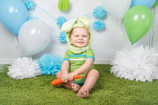 Portret van leuke schattige Kaukasische babyjongen in vakantie Pasen konijn kostuum met grote oren, gekleed in groene kleren onesie, zittend op zachte pluizige tapijt tapijt in studio, rode wortel te houden — Stockfoto