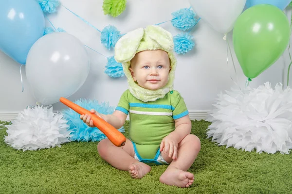 Portrait of cute adorable Caucasian baby boy in holiday Easter bunny rabbit costume with large ears,  dressed in green clothes onesie, sitting on soft fluffy rug carpet in studio, holding red carrot — Stock Photo, Image