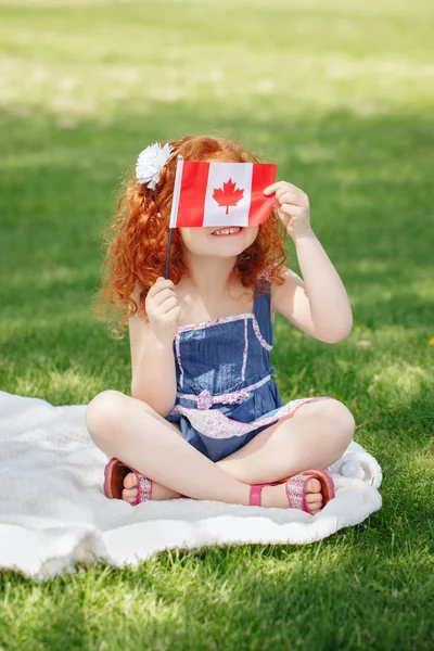 Potret anak kecil berambut merah yang lucu memegang bendera Kanada dengan daun maple merah, duduk di rumput di taman di luar, merayakan Hari Kanada — Stok Foto
