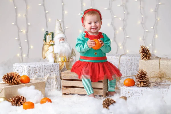 Potret gadis pirang kaukasia dengan mata biru dengan kostum elf merayakan Natal atau liburan Tahun Baru. Smilingaby duduk dengan buah jeruk keprok clementine — Stok Foto