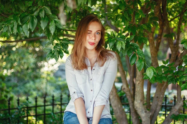特写肖像美丽微笑的白种白种女孩长着金色的头发和蓝色的眼睛穿着白色的衬衫和牛仔裤在夏天公园外面在绿色的叶子树在照相机看 — 图库照片