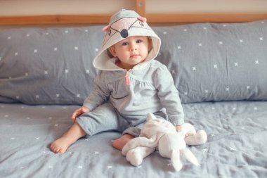 Portre sevimli sevimli Beyaz sarışın gülümseyen kız bebek yatak yatak odasında oturma ve tutmak oyuncak tilki kedi hayvan kukuleta ile gri pijama içinde mavi gözlü. Mutlu çocukluk yaşam tarzı kavramı