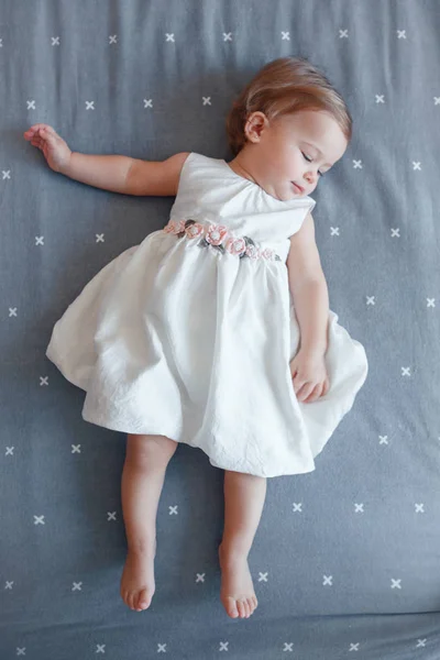 一岁穿着白色连衣裙的可爱迷人的白种金发女婴的肖像躺在卧室的灰色床单上 从上面看 快乐的童年生活理念 — 图库照片