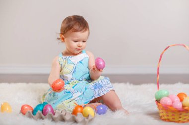 Mavi elbiseli Studio beyaz yumuşak tüylü halı halı üzerinde oturan sevimli sevimli beyaz bebek kız. Geleneksel kutsal Hıristiyan tatil kutlayan Paskalya renkli yumurta ile oynayan çocuk çocuk.