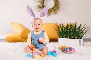Mor tavşan kulakları stüdyoda oturan giymiş kot elbise sevimli sevimli beyaz bebek kız. Geleneksel kutsal Hıristiyan tatil kutlayan Paskalya renkli yumurta ile oynayan çocuk çocuk.
