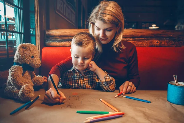 Portrait of young Caucasian mother and son sitting in restaurant drawing with colored pencils on craft brown paper. Family together in cafe. Lifestyle authentic real people. Toned with filters.