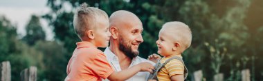 Oğulları yazın parkta olan beyaz bir baba. Çocukların babası kucağında. Otantik yaşam tarzı dokunaklı bir an. Aile hayatı mutlu babalık. Web sayfası başlığı.