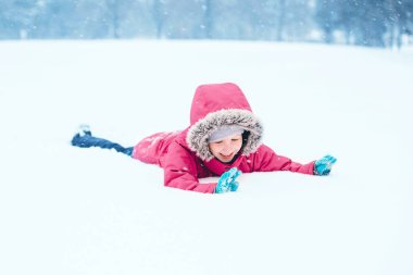 Tatlı, komik, beyaz, gülümseyen, sıcak elbiseli, pembe ceketli, kar yağışlı soğuk kış günlerinde yerde karla oynayan kız çocuğu. Çocuklar açık havada mevsimlik aktiviteler.