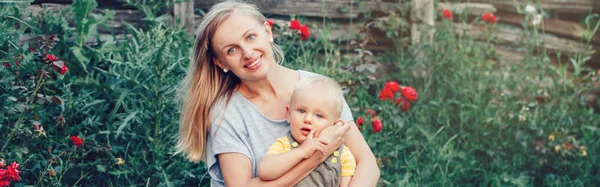 Caucasian Ukrainian mother with long blonde hair holding toddler baby boy on her lap knees on summer day in park. Happy motherhood parenting lifestyle. Web banner header for website.