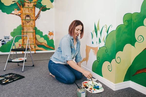 Lifestyle creative hobby and freelance artistic work side job concept. Caucasian woman artist hand painting murals on walls indoor at apartment or studio school with acrylic paints.