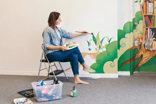 Lifestyle creative hobby and freelance artistic work side job concept. Caucasian woman artist hand painting murals on walls indoor at apartment or studio school with acrylic paints.