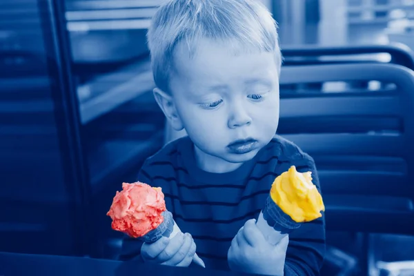 Anak lucu kaukasia yang lucu memegang dua es krim berwarna kuning merah di wafel cone. Anak itu mencoba memilih makanan. Bingung, ekspresi wajah emosional. Dada dengan warna klasik tahun 2020 biru . — Stok Foto