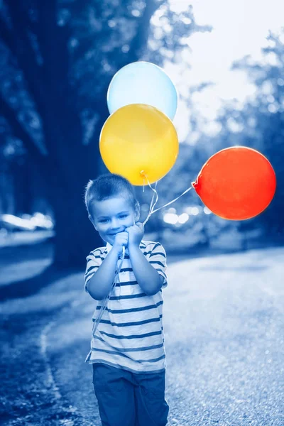 Niño caucásico niño pequeño con globos de colores en el parque al aire libre. Al chico le gusta jugar. Feliz fiesta de cumpleaños. Auténtica infancia estilo de vida. Tonificado con el clásico color azul 2020 . — Foto de Stock