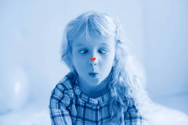 Смешная весёлая белая милая очаровательная девочка смотрит на свой нос с наклейкой на сердце. Косоглазый пацан щурящийся. День святого Валентина праздник концепция. Тонированные классическим синим цветом 2020 года . — стоковое фото