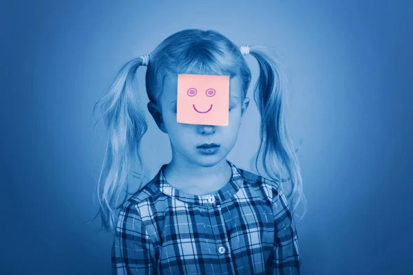 Sad unhappy Caucasian preschool girl with funny pink sticky note paper on her face. Kid expressing emotions. Concept of difficulties, problems of childhood. Toned with classic blue 2020 color. — Stock Photo, Image