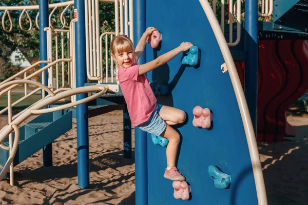 Feliz sorrindo pequena menina pré-escolar escalando parede de rocha no playgr — Fotografia de Stock