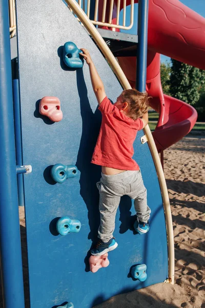 Kis óvodás fiú piros pólóban sziklát mászik a Playgr-ben. — Stock Fotó