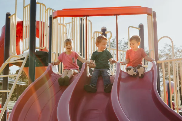 Ativo crianças caucasianas felizes deslizando no pátio do parque infantil — Fotografia de Stock