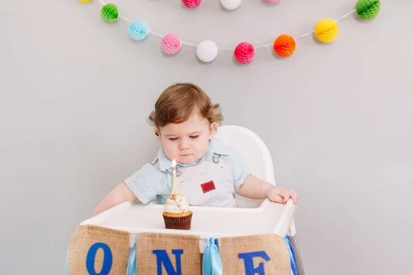 Schattig schattig blank jongetje dat zijn eerste verjaardag thuis viert. Kind peuter zit in kinderstoel smakelijk cupcake dessert te eten met één kaars. Gelukkige verjaardag concept. — Stockfoto