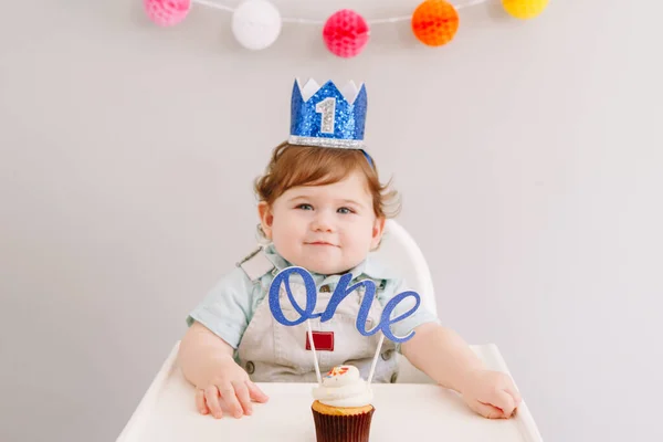Roztomilý rozkošný kavkazský chlapeček v modré koruně, oslavující své první narozeniny doma. Dítě dítě batole sedí na vysoké židli jíst chutné dortík dezert se slovem topper. Happy birthday concept. — Stock fotografie