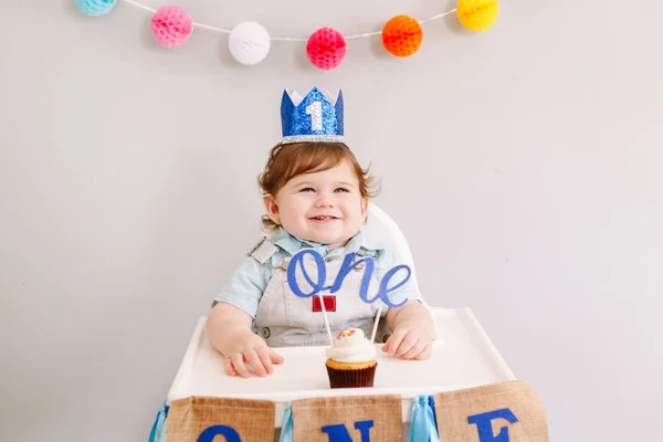 Bonito menino caucasiano adorável em coroa azul celebrando seu primeiro aniversário em casa. Criança criança criança sentada em cadeira alta comendo sobremesa de cupcake saboroso com topper palavra. Conceito de feliz aniversário . — Fotografia de Stock