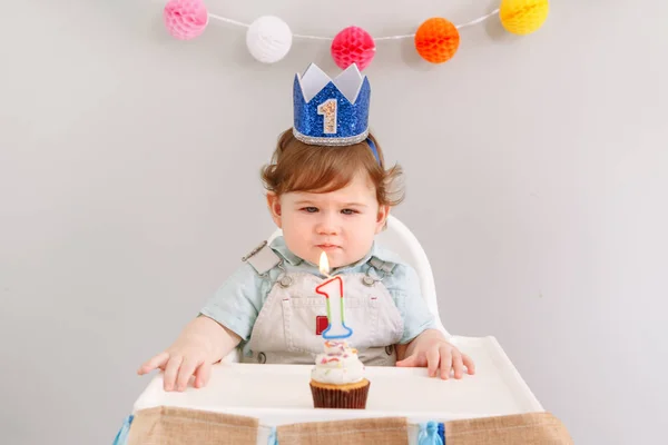 Cute adorable Caucasian baby boy in blue crown celebrating his first birthday at home. Child kid toddler sitting in high chair looking at cupcake dessert with lit candle. Happy birthday concept. — Stock Photo, Image