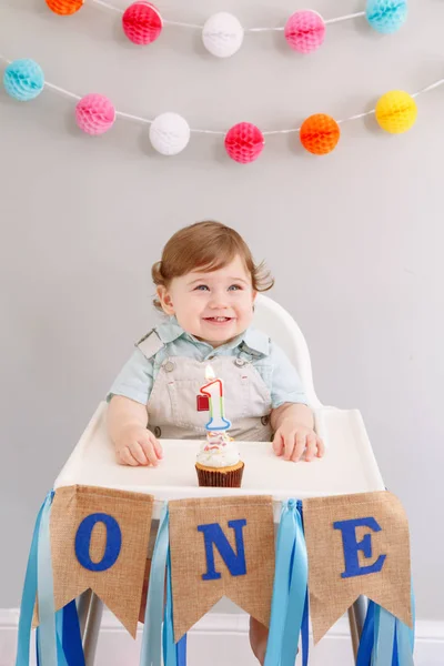 Happy smiling cute Caucasian baby boy celebrating his first birthday at home. Child kid toddler sitting in high chair eating tasty cupcake dessert with lit candle. Happy birthday concept. — ストック写真