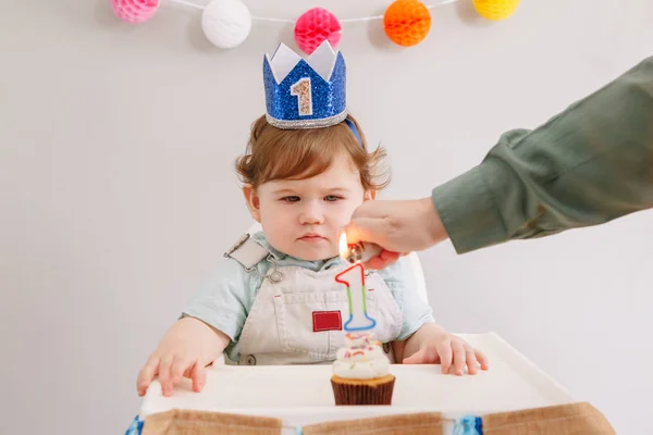 Bayi laki-laki Kaukasia imut bermahkota biru merayakan ulang tahun pertamanya di rumah. Anak balita duduk di kursi tinggi melihat makanan penutup cupcake. Ibu menyalakan lilin ulang tahun pada kue . — Stok Foto