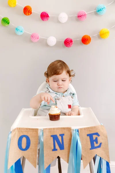 Bayi laki-laki Kaukasia yang manis dan sedih merayakan ulang tahun pertamanya di rumah. Anakkecilbalita duduk di meja tinggi makan makanan penutup cupcake lezat. Selamat ulang tahun konsep gaya hidup. — Stok Foto