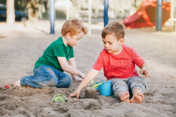 Dos niños caucásicos sentados en arenero jugando con juguetes de playa. Niños amigos divirtiéndose juntos en el patio de recreo. Actividad al aire libre de verano para niños. Tiempo libre estilo de vida infancia . — Foto de Stock