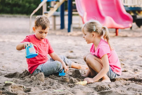 Två vita barn sitter i sandlåda och leker med strandleksaker. Flicka och pojkvänner har kul på lekplatsen. Sommar utomhus aktivitet för barn. Fritid livsstil barndom. — Stockfoto