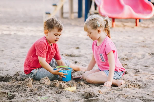 Dos niños caucásicos sentados en arenero jugando con juguetes de playa. Niña y niños amigos divirtiéndose juntos en el patio de recreo. Actividad al aire libre de verano para niños. Tiempo libre estilo de vida infancia . — Foto de Stock