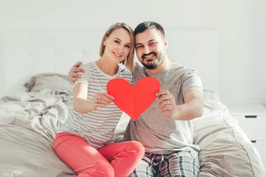 Happy strong marriage. Happy funny couple in love holding red paper heart. Heterosexual family man and woman sitting on bed in bedroom at home hugging. Real people authentic lifestyle. 