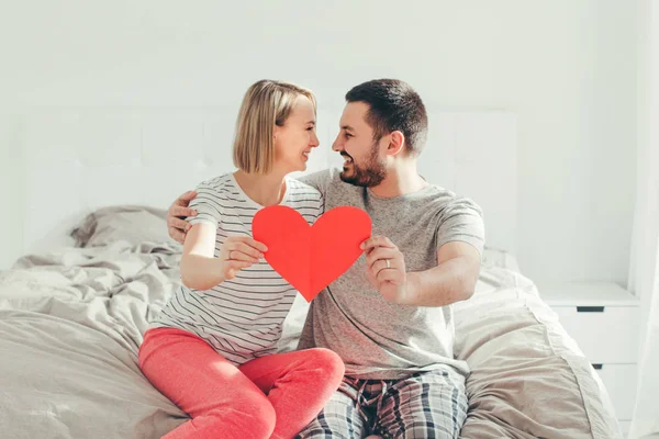 Happy strong marriage. Happy funny couple in love holding red paper heart. Heterosexual family man and woman sitting on bed in bedroom at home hugging. Real people authentic lifestyle. — 图库照片
