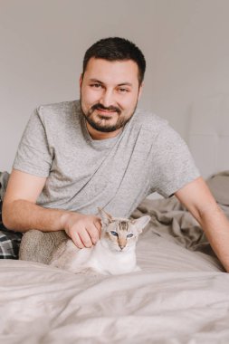 Mutlu, beyaz, gülümseyen sakallı bir adam evdeki yatak odasında oturmuş mavi gözlü, oryantal noktalı kediyi okşuyor. Evcil hayvanı olan gururlu bir hayvan sahibi.. 