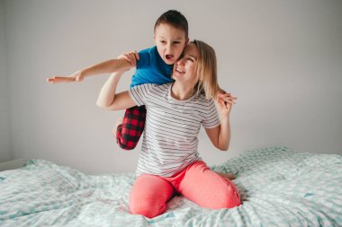 Gülümseyen beyaz anne ve oğlu evdeki yatak odasında oynuyorlar. Çocuk annesinin omuzlarında oturuyor ve gülüyor. Aile birlikte eğleniyor. Mutlu çocukluk samimi, otantik yaşam tarzı.
