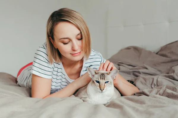 骄傲的宠物主人快乐的白种人微笑的金发女人爱抚着蓝眼睛的东方猫 漂亮的女孩躺在家里的卧室里爱抚家养的猫 — 图库照片