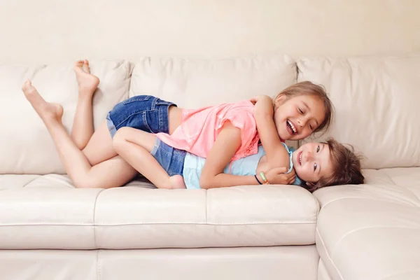 かわいい白人の二人の女の子が家で遊んでいます 愛らしい子供たちが一緒にソファで抱き合って横たわっています 本物の率直なライフスタイル国内生活の瞬間 幸せな友人姉妹関係 — ストック写真