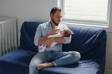 Orta yaşlı beyaz bir baba yeni doğmuş bebeği şişe sütle besliyor. Sallanan çocuğu elinde tutan bir ebeveyn. Otantik yaşam tarzı gerçek bir an. Bekar baba aile hayatı konsepti..