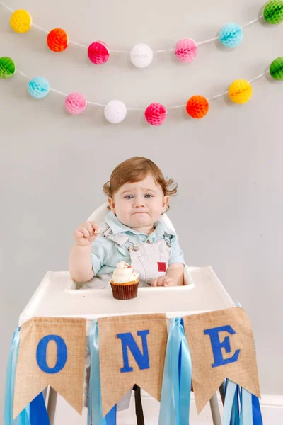 かわいい愛らしい悲しい動揺白人の赤ちゃん男の子自宅で彼の最初の誕生日を祝う 高椅子テーブルに座っている幼児の幼児はおいしいカップケーキデザートを食べています 幸せな誕生日のライフスタイルのコンセプト — ストック写真