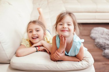 Evde oynayan iki küçük şirin beyaz kız kardeş var. Sevimli gülümseyen çocuklar kanepede birlikte yatıyorlar. Otantik samimi yaşam tarzı ev hayatı anı. Mutlu arkadaşlar kız kardeşler ilişkisi. 