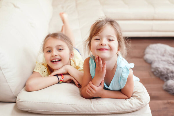 Two cute little Caucasian girls siblings playing at home. Adorable smiling children kids lying on couch together. Authentic candid lifestyle domestic life moment. Happy friends sisters relationship. 