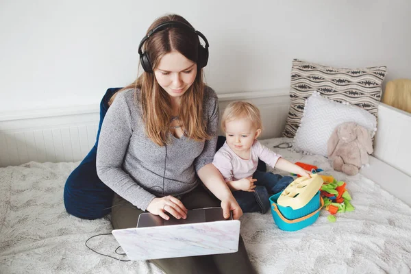 Young Caucasian mother blogger with baby working on laptop from home. Workplace of freelance woman student with kid toddler. Stay at home single mom earning money at online job.