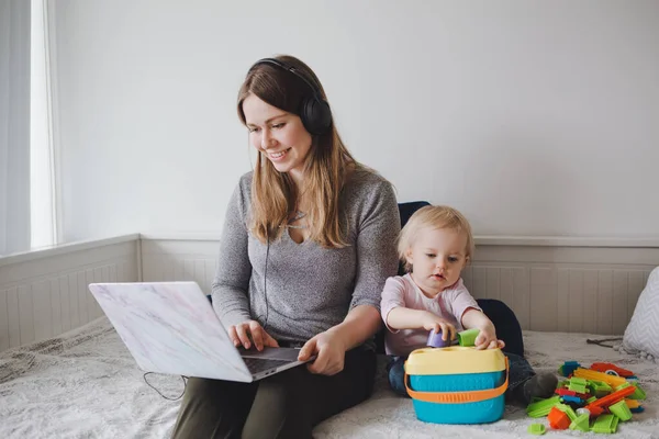 Young Caucasian mother blogger with baby working on laptop from home. Workplace of freelance woman student with kid toddler. Stay at home single mom earning money at online job.