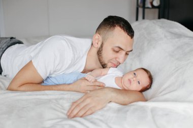 Beyaz bir baba yeni doğmuş bir bebekle yatak odasında yatıyor. Erkek ebeveyn kucaklaşması, kız evlat kucaklaması. Otantik yaşam tarzı samimi anı. Gururlu genç baba. Aile Babaları Günü.