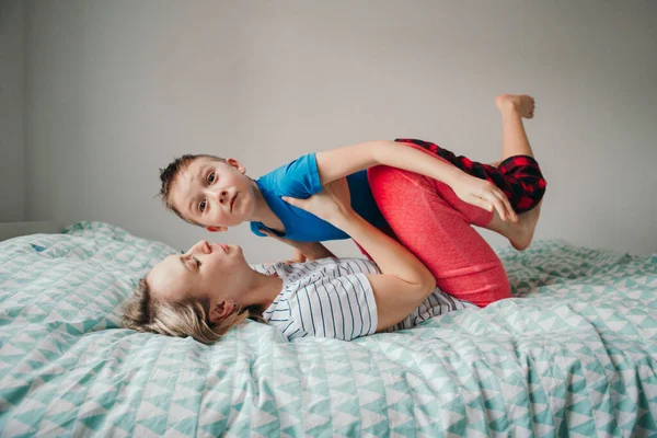 自宅の寝室で遊んでいる白人の母親と少年の息子 膝の上で母親が子供を揺らす 家族みんなで楽しんでる 幸せな率直な本格的な子供時代のライフスタイル — ストック写真