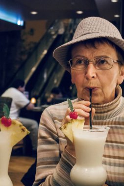 Güzel, şık, yaşlı bir kadın barda tatlı kokteyl içiyor. Şapkalı ve bardaklı yaşlı beyaz kadın restoranda içkisini yudumluyor ve hayatın tadını çıkarıyor. Mutlu olgunluk çağı konsepti.